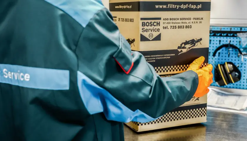 Pracownik w kombinezonie i rękawiczkach trzyma karton z logo Bosch Service.