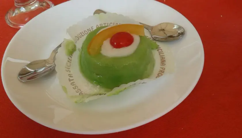 Zielone lody cassate, ozdobione wiśnią i skórką pomarańczy, na białym talerzyku z łyżeczką.
