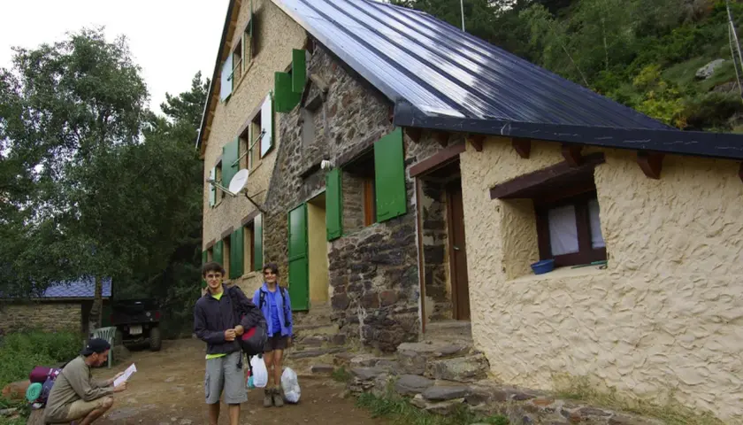 Llegada al refugio de Vallferrera. Dos jóvenes con mochilas y un hombre sentado revisando un mapa esperan en la entrada de piedra y estuco.
