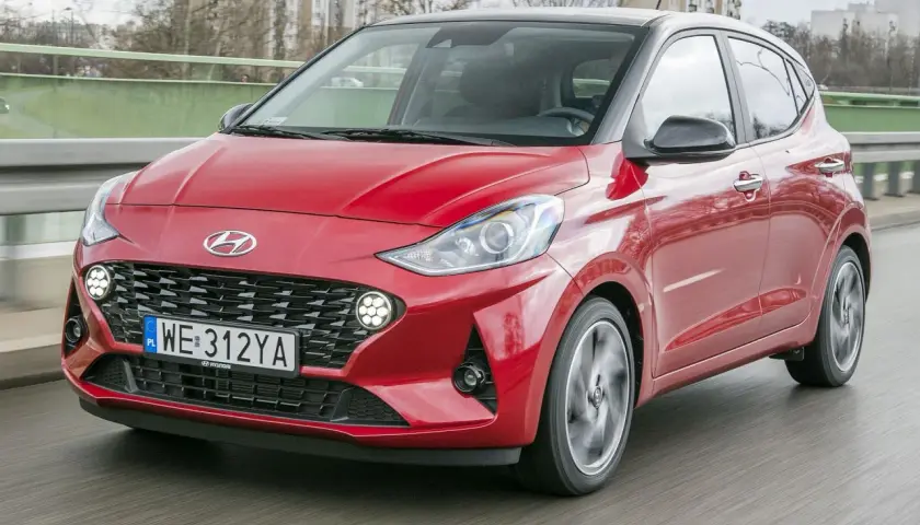 Czerwony Hyundai i10 z czarnym dachem pędzi po drodze. W tle widać budynki i drzewa.