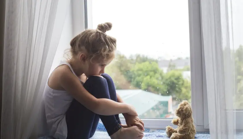Smutna dziewczynka siedzi przy oknie, przytulając kolana. Obok niej pluszowy miś. Obraz symbolizuje wykluczenie z grupy rówieśniczej i poczucie samotności.