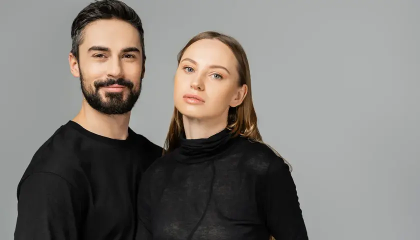 Para, która wygląda jakby mogła nosić ubrania unisex. Mężczyzna z brodą i kobieta z długimi włosami, oboje w czarnych, prostych ubraniach.