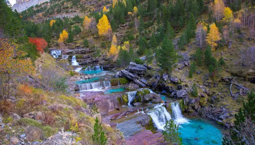 Cascada en las gradas de soaso, con aguas turquesas y rocas rojizas, rodeada de árboles otoñales y pinos verdes.