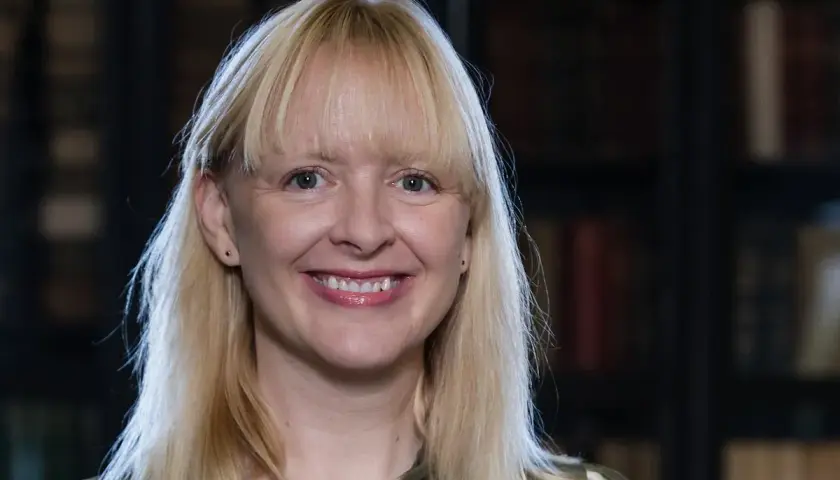 Uśmiechnięta blondynka z grzywką, w tle biblioteka. Jej spojrzenie sugeruje, jakie są relacje między autorem a jego literackim alter ego – pełne refleksji.
