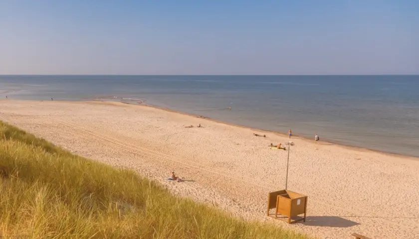 Słoneczna plaża w Kuźnicy nad morzem. Kilka osób odpoczywa na piasku, w oddali widać spokojne morze i błękitne niebo.