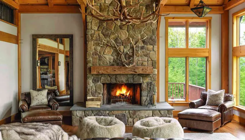 A cozy living room with a stone fireplace, antler chandelier, and leather armchairs embodies traditional rustic interior design.