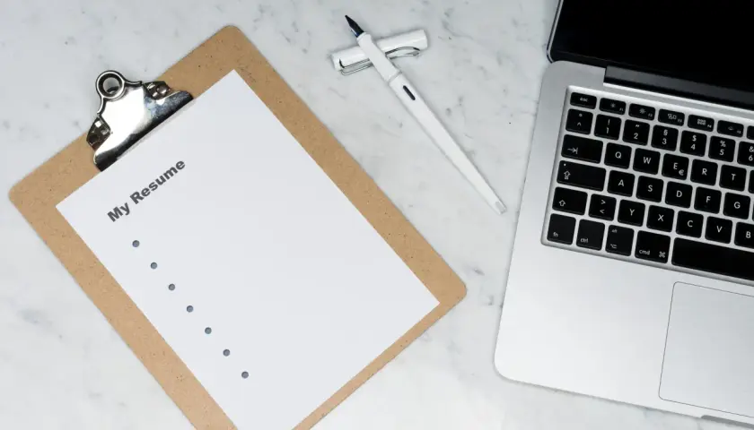 Na biurku leży laptop, białe pióro i kartka z napisem "My Resume". Idealne miejsce do przygotowania CV po angielsku.