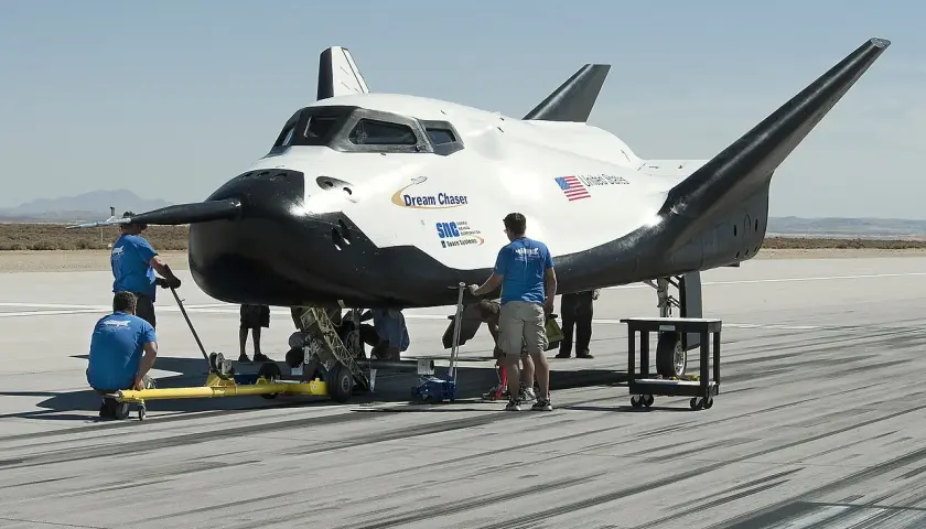 Dream Chaser, kosmiczny pojazd, przygotowywany do startu. Zespół techników pracuje przy jego podwoziu na pustynnym lotnisku.