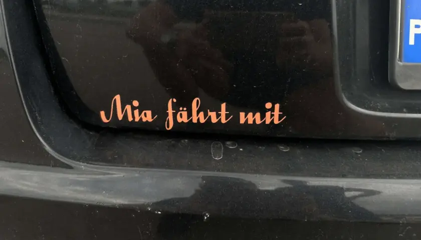 Pomarańczowa naklejka na czarnym lakierze samochodu z napisem "Mia fährt mit". Chcesz wiedzieć, jak usunąć naklejki z samochodu?