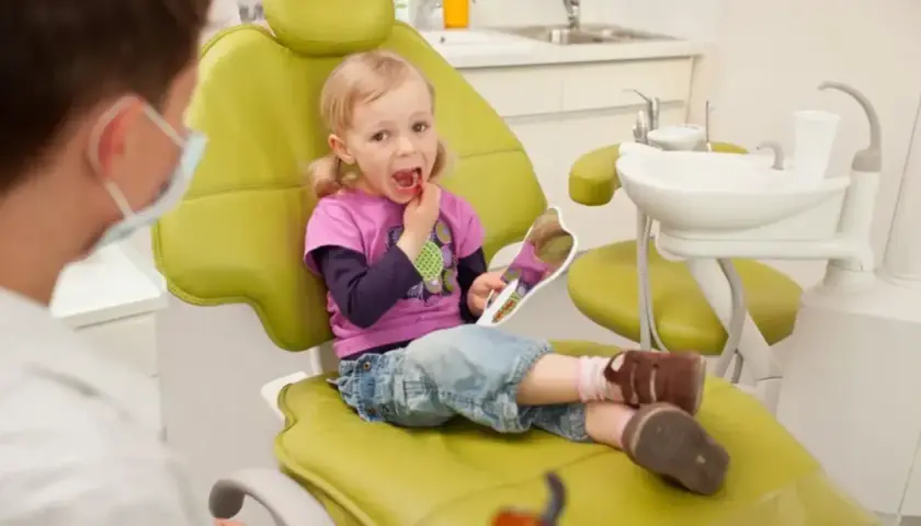 Mała dziewczynka z lusterkiem sprawdza swoje zdrowe zęby mleczne podczas wizyty u dentysty.