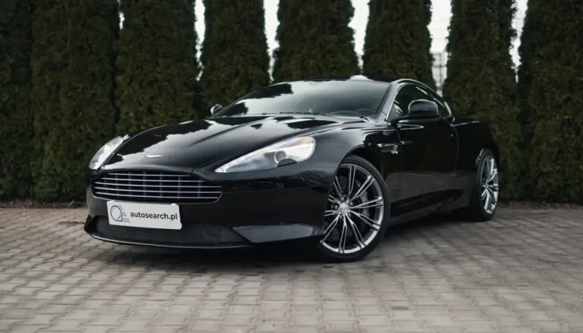 Czarny Aston Martin DB9 lśni na tle zielonych żywotników. Elegancki, sportowy samochód z charakterystycznym grillem i felgami.