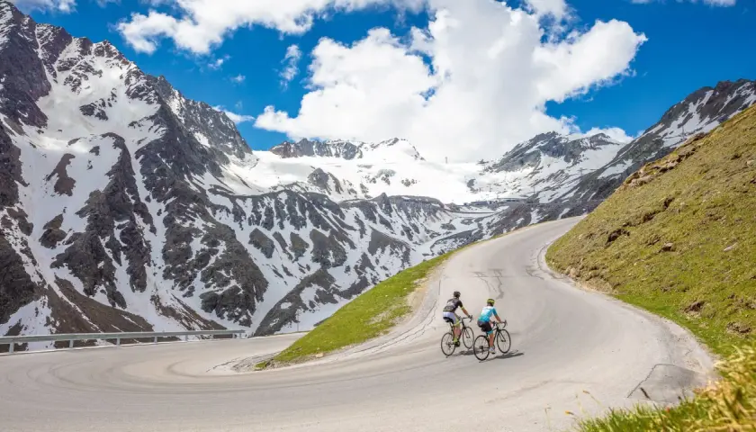 Two cyclists enjoy an epic adventure on a winding road, part of amazing austria bike tours, with snow-capped mountains in the background.