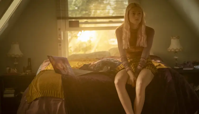Una ragazza bionda con un vestito a quadri siede sul letto, illuminata dalla luce del tramonto. Una scena che fa pensare a una recensione di Euphoria.
