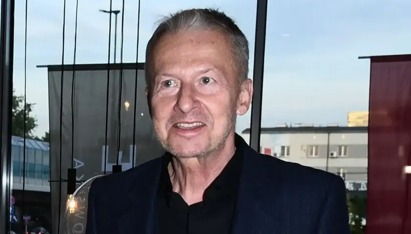 Bogusław Linda w granatowej marynarce i czarnej koszuli, uśmiecha się.