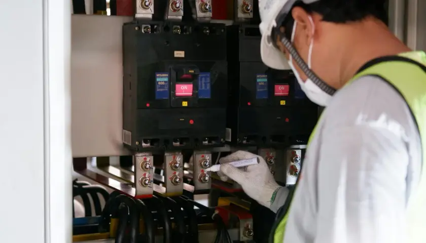 Elektryk w białym kasku i masce zabezpieczającej oznacza śruby przy głównym bezpieczniku prądu.