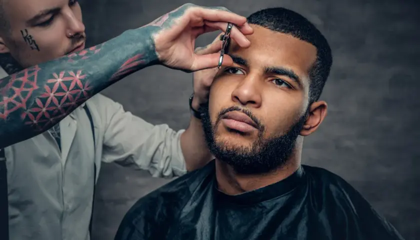 Barbero con tatuajes en el brazo recorta las cejas a un hombre con un corte buzz cut y barba.