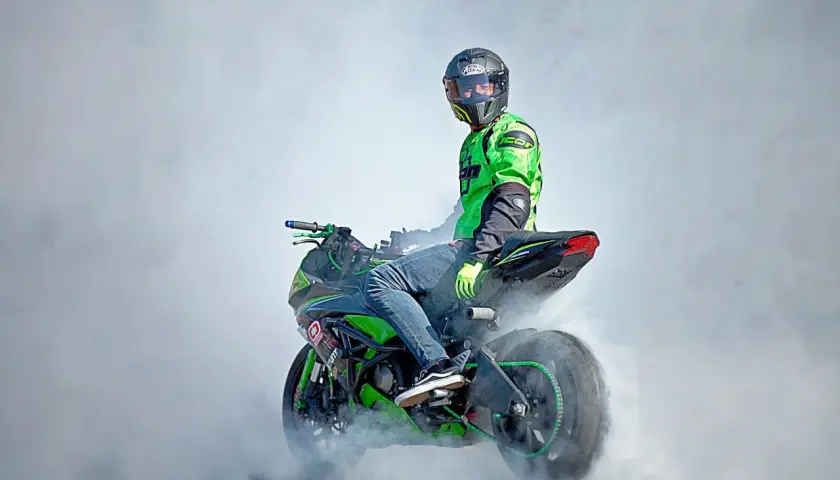 Motociclista con casco y traje verde realiza un caballito, generando humo. La moto, con detalles verdes, parece lista para una demostración de homologación e24.