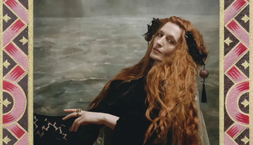 Florence Welch, jak królowa, z rudymi włosami i w czarnej sukni, na tle morskiego krajobrazu.