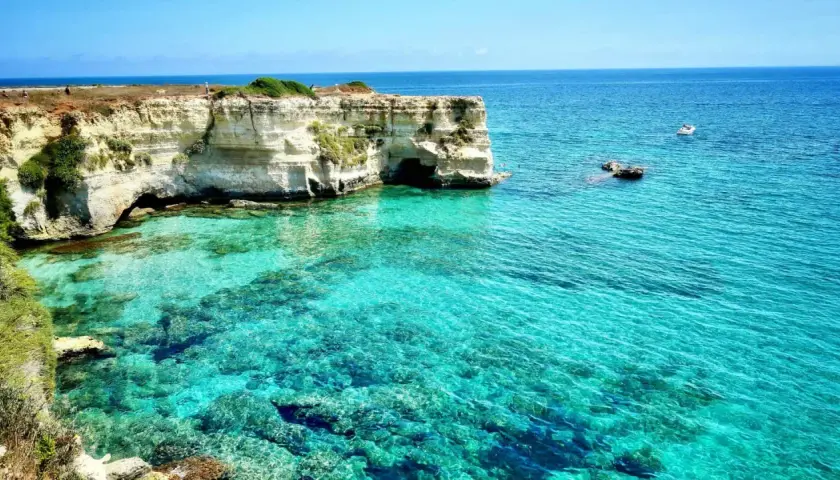 Turquoise water laps against white cliffs with caves, a boat sails in the distance.
