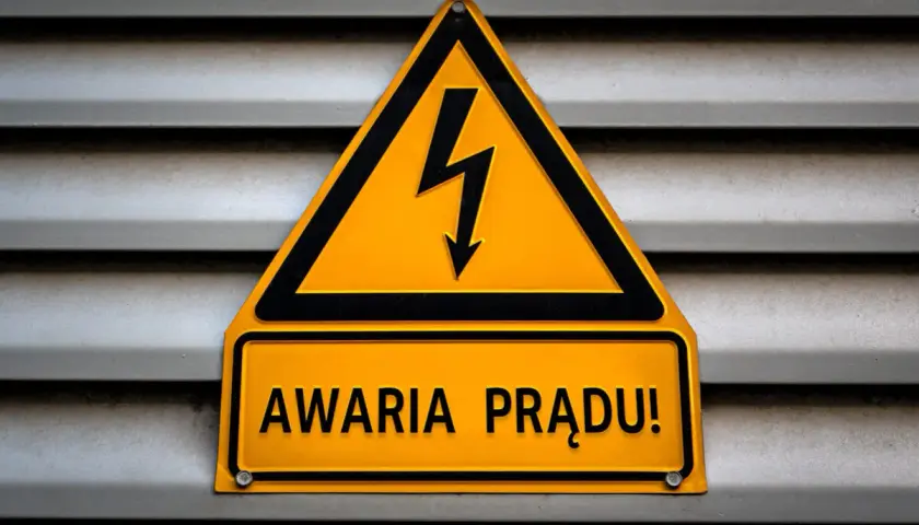 Żółty trójkąt z symbolem pioruna i tabliczka "AWARIA PRĄDU!" na szarej, metalowej powierzchni.