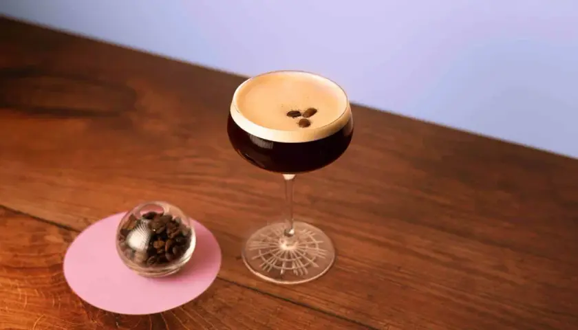 Espresso Martini with coffee beans on top, next to a small glass of coffee beans on a pink coaster.