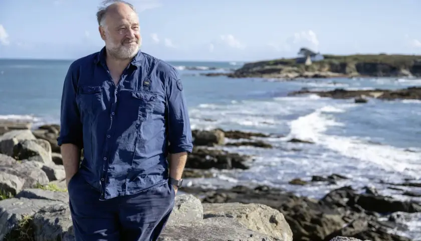 Ein Mann in blauer Kleidung steht am Meer. Die bannalec reihenfolge der Wellen spiegelt sich in seiner entspannten Haltung wider.
