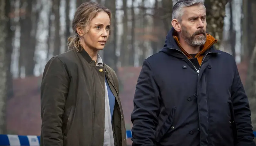 Två personer, en kvinna och en man, står i en skog. Bakom dem syns blåvit avspärrningsband. Kanske är det Sandor Berg som utreder ett brott.