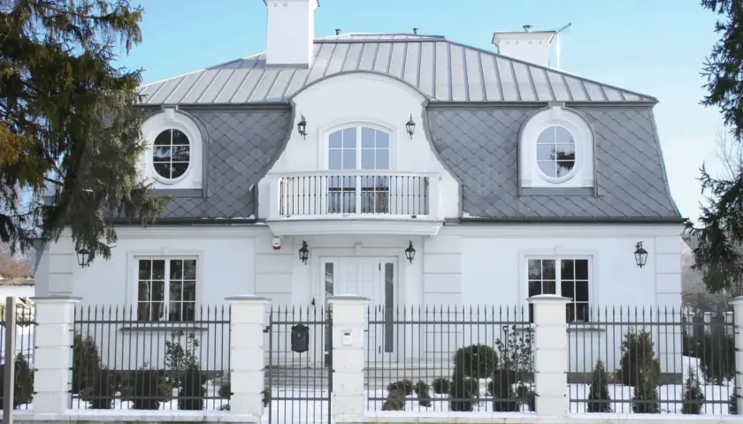 Elegancki dom z białym tynkiem i szarym, łamanym dachem mansardowym. Okna okrągłe i prostokątne, balkon nad wejściem.