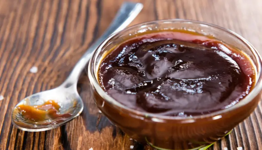 Un cuenco de cristal lleno de deliciosa salsa barbacoa casera, con una cuchara al lado y romero fresco.
