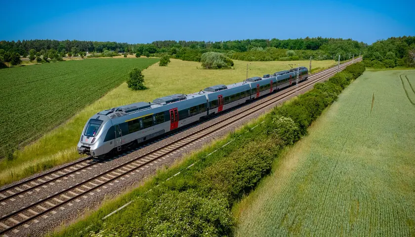 Moderner Triebzug fährt auf einer **railway deutsch** durch grüne Felder und Wälder unter blauem Himmel.
