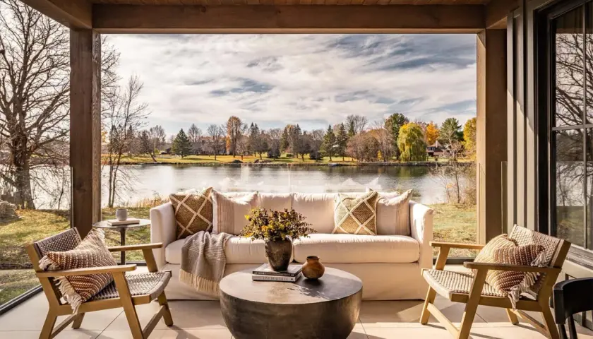 Cozy lakehouse interior design with a white sofa, chairs, and coffee table on a covered patio overlooking a serene lake and autumn foliage.