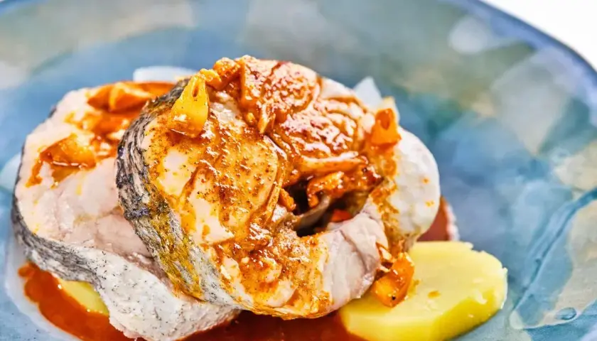 Rodajas de pescado blanco cubiertas con una sabrosa salsa gallega, servidas sobre patatas cocidas en un plato azul.