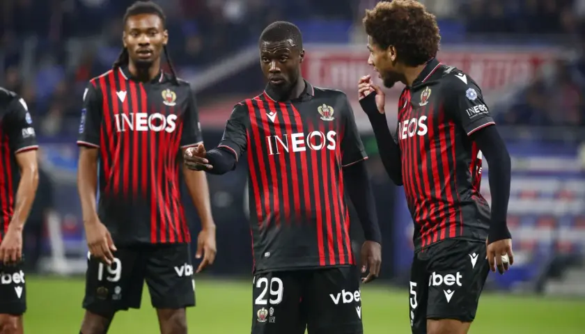 Piłkarze OGC Nice w czarno-czerwonych strojach dyskutują na boisku. Jeden z nich, z numerem 29, wskazuje palcem.