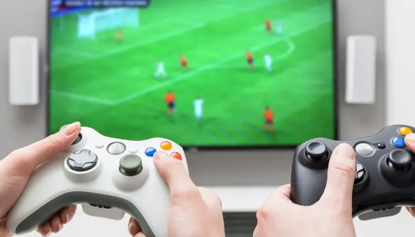 Dwie osoby grają w gry sportowe na konsoli, trzymając kontrolery przed ekranem telewizora z boiskiem piłkarskim.