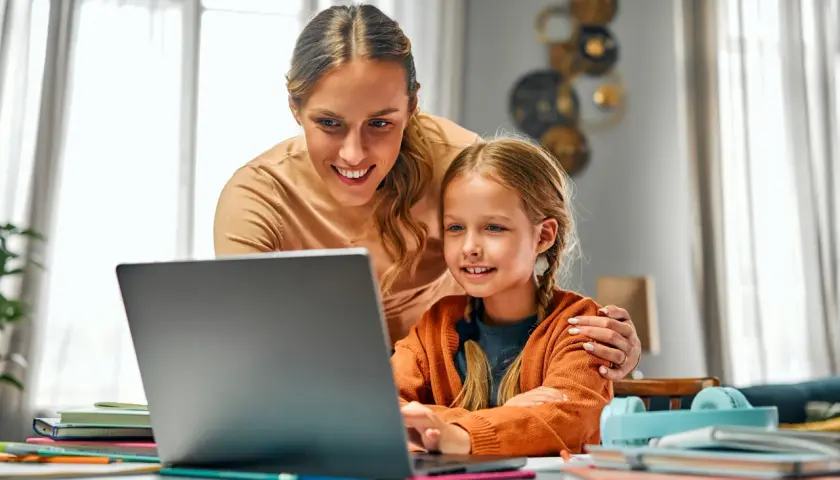 Mama wspiera córkę w nauce przy laptopie. To piękny przykład opieki nad zdrowym dzieckiem, łączącej edukację z bliskością.