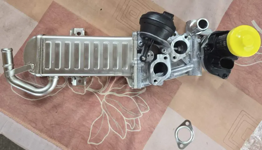 VW AGR-Kühler mit schwarzem Stellmotor und gelber Kappe auf gemustertem Untergrund.