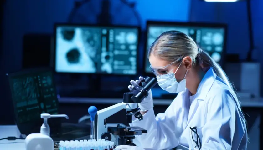 Diagnosta laboratoryjny w okularach i masce bada próbki pod mikroskopem. Na ekranach komputera widać wyniki badań.