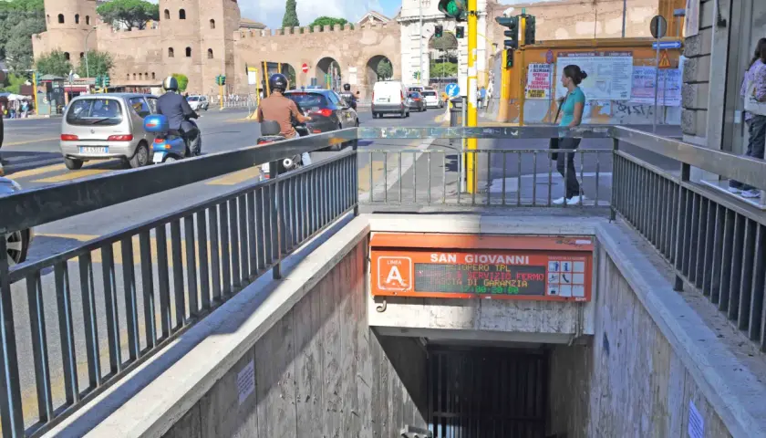 Ingresso metro San Giovanni, collegamento metro A. Traffico intenso con auto e scooter, sullo sfondo le antiche mura romane.