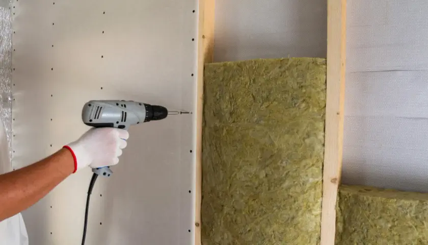 Installazione di pannelli isolanti per un maggiore comfort acustico. Una mano con guanto bianco avvita un pannello di cartongesso su una struttura in legno con isolamento.