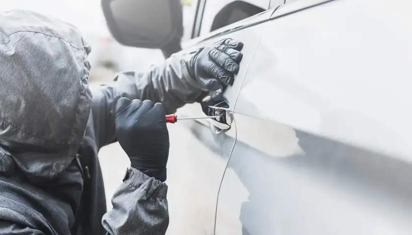 Osoba w kapturze i rękawiczkach próbuje otworzyć auto śrubokrętem. Sen o kradzieży auta może symbolizować utratę kontroli.