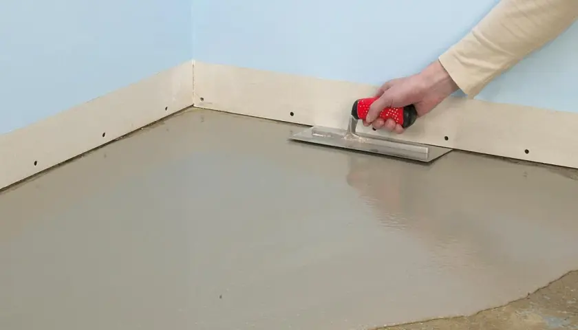 En hand spacklar golv med en bredspackel. Det nya golvet är jämnt och grått.