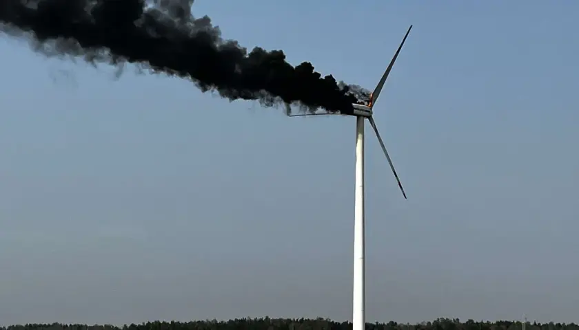 Płonąca turbina wiatrowa z gęstym, czarnym dymem unoszącym się na tle błękitnego nieba.