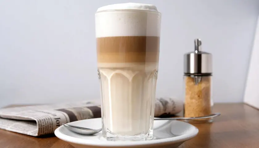 Ein geschichteter Latte Macchiato im Glas, daneben eine Zeitung und ein Zuckerspender. Milchkaffee vs. Latte Macchiato – die Wahl ist schwer!