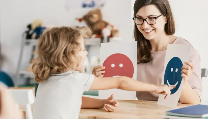 Dziecko wskazuje na uśmiechniętą buźkę, rozwijając inteligencję intrapersonalną. Terapeuta z uśmiechem prezentuje karty emocji.