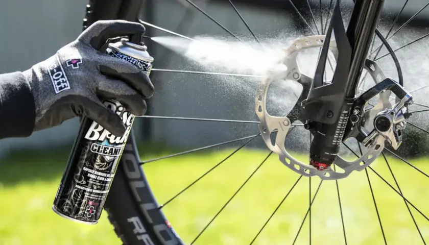 Gloved hand sprays Muc-Off Disc Brake Cleaner onto a bike's rotor, demonstrating how to clean up brake fluid for optimal performance.