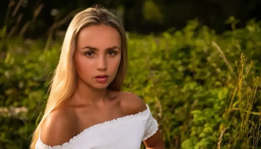 Portret w plenerze młodej kobiety z długimi blond włosami, w białej bluzce z odkrytymi ramionami, na tle zielonej roślinności.