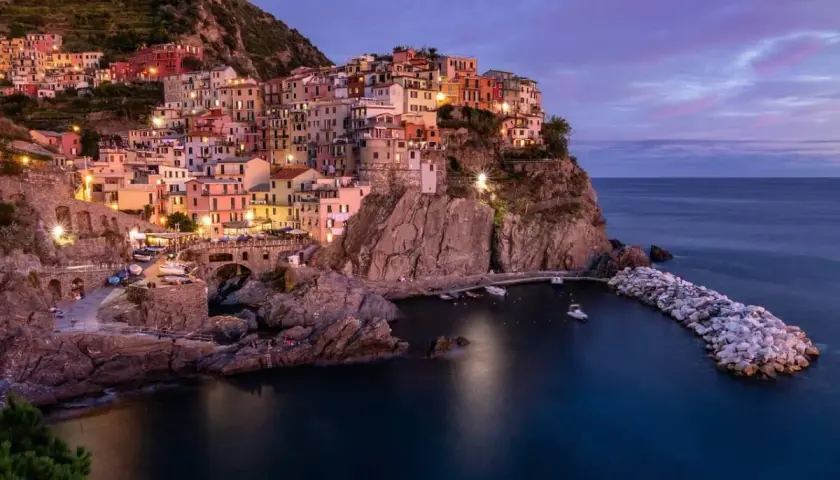 Manarola, una delle perle della strada panoramica 5 Terre, si illumina al crepuscolo. Case colorate aggrappate alla roccia, un piccolo porto e il mare blu intenso.