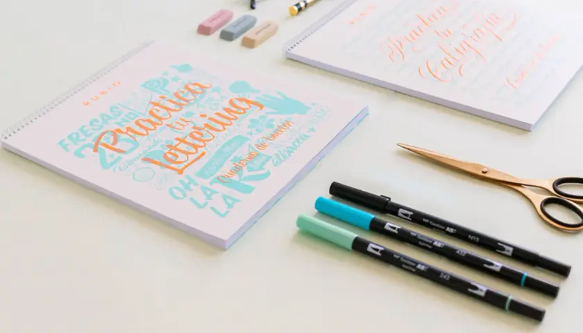 Cuadernos de "Practica Lettering" y "Practica tu Caligrafía" con rotuladores y tijeras doradas.