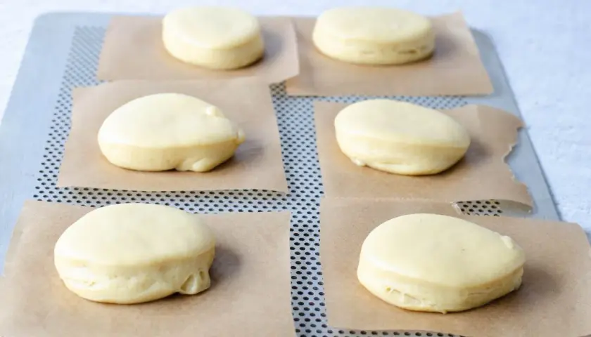 Seis bolas de fraile crudas, listas para hornear, descansan sobre papel de horno en una bandeja metálica.
