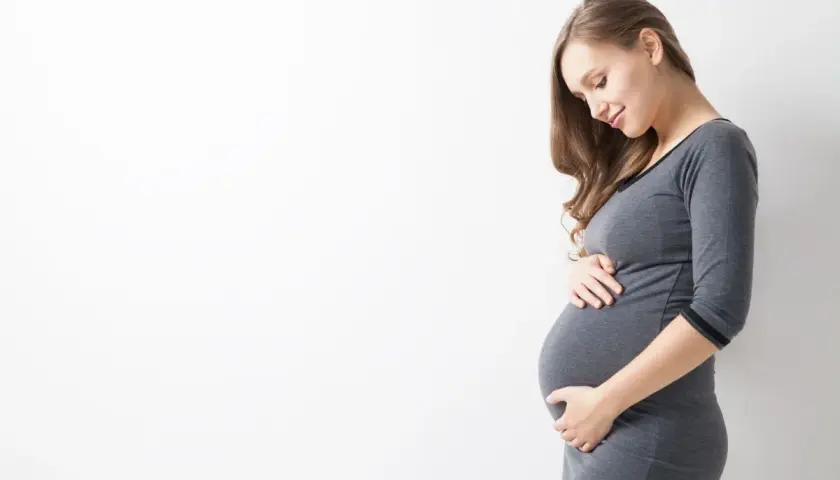 Uśmiechnięta ciężarna kobieta w szarym ubraniu delikatnie dotyka brzucha. To naturalne zmiany, jak rozstępy w ciąży, które są częścią tej wyjątkowej podróży.